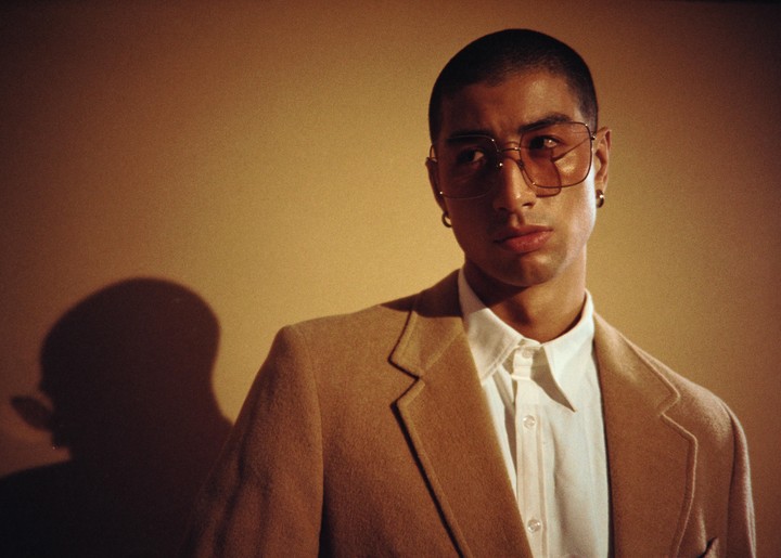 A man wearing a tan jacket and glasses looks to the side, with a shadow of his head visible on the wall behind him.