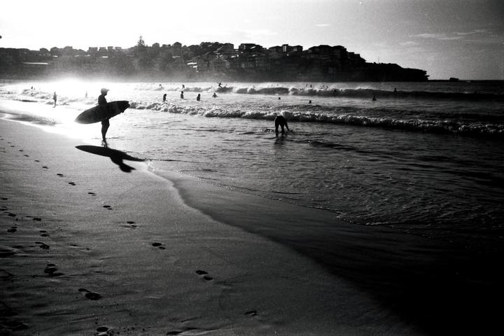 A surfer stands on a beach with a surfboard, looking out at the ocean with waves and other people in the water, with a town on a hill in the background.