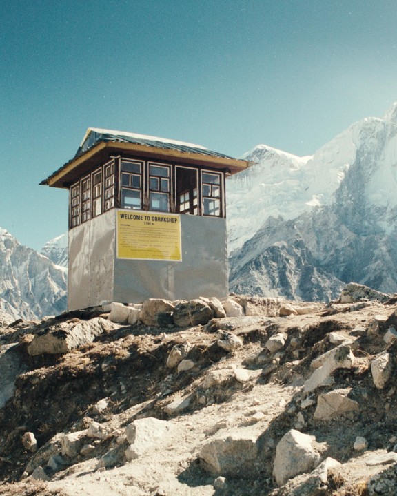 A small building with a yellow sign that reads "WELCOME TO GORAKSHEP" stands on a rocky hill with snow-capped mountains in the background.