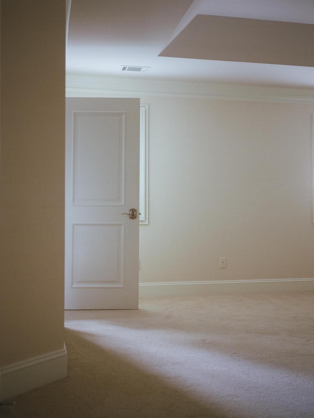 A partially open white door reveals a room with light-colored walls and carpet, with a shadow cast across the floor.