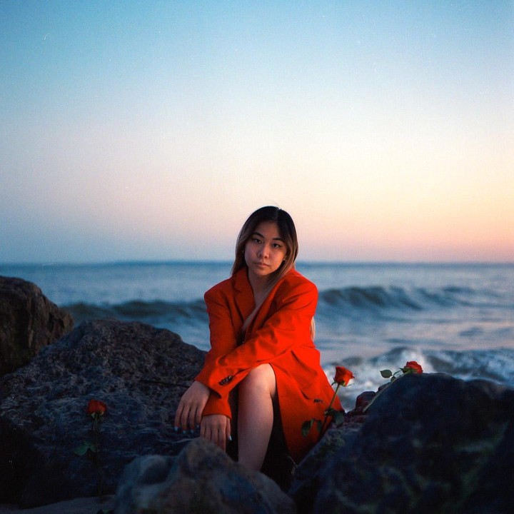 A woman in a red jacket sits on rocks by the ocean at dusk, with red roses scattered around her.