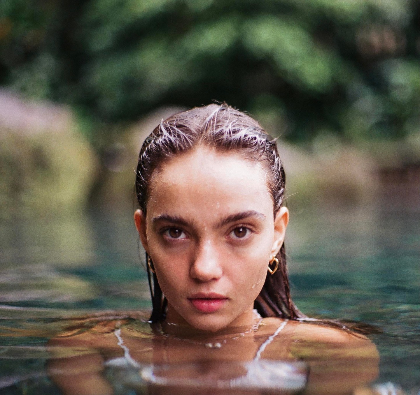 A woman with wet hair and a heart earring looks directly at the viewer from a natural spring.