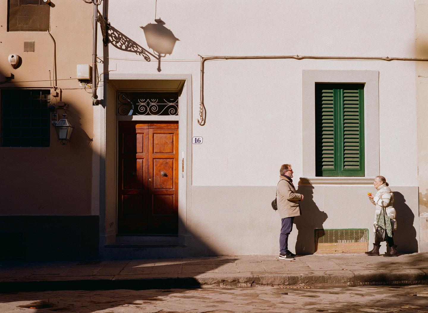 A man and a woman stand on a sunlit street next to a building with a wooden door and a window with green shutters.