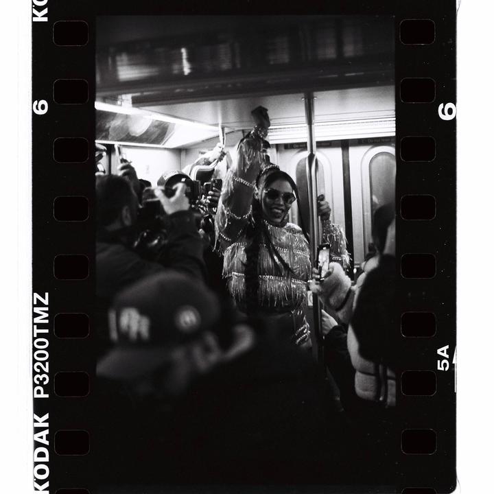 A woman in sunglasses and a fringed jacket smiles while holding onto a pole in a crowded subway car.