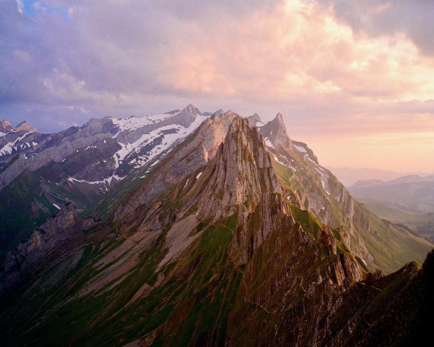 A mountain range with snow-capped peaks and green slopes is illuminated by the golden hour light under a cloudy sky.