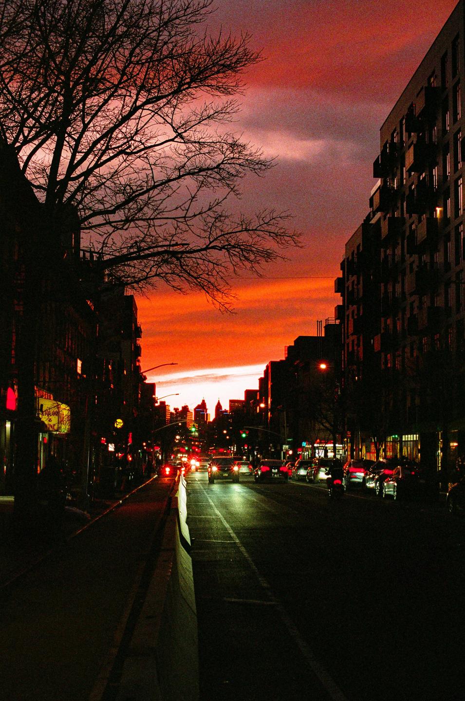 A street scene at sunset shows buildings and a bare tree silhouetted against a deep red and orange sky, with car lights visible on the road.