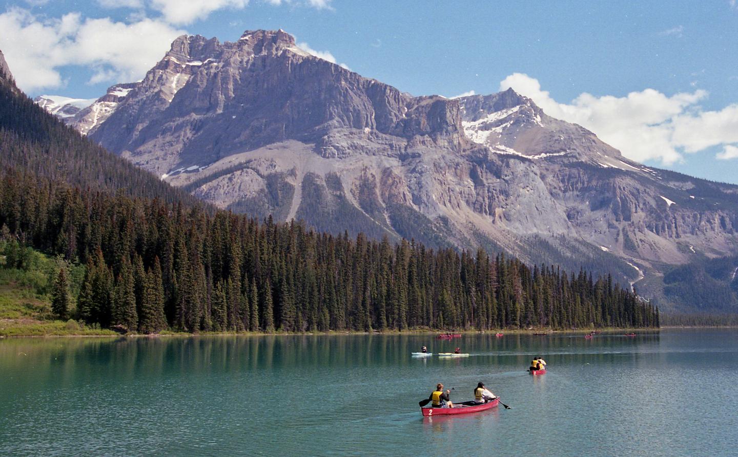 Several people in canoes and kayaks paddle on a lake surrounded by a dense forest and a large mountain range.