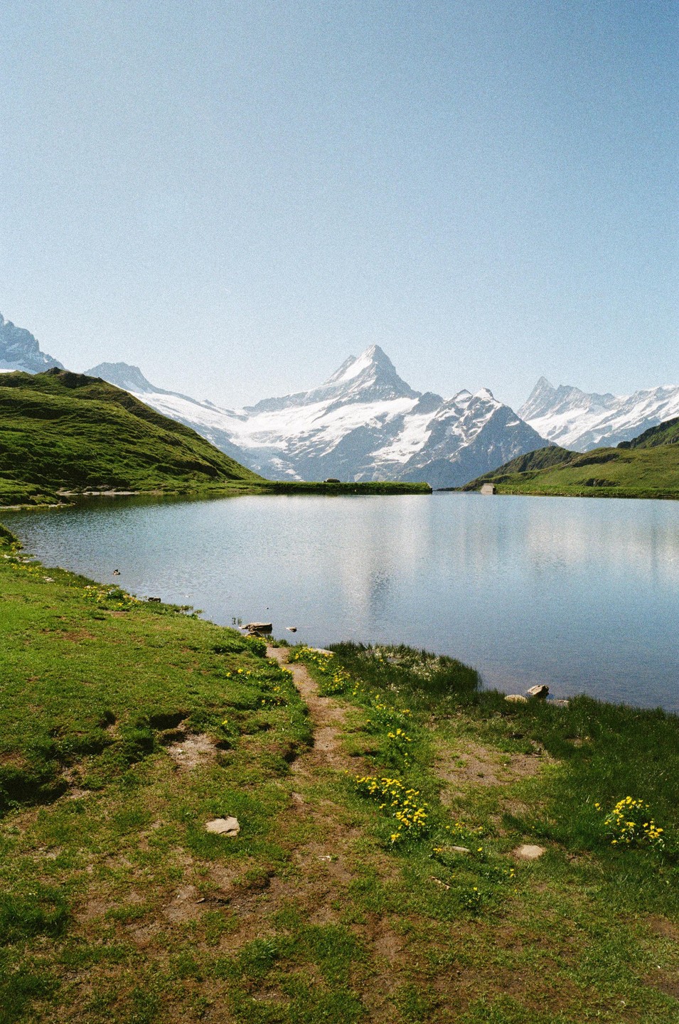 A calm lake reflects snow-capped mountains under a clear sky, with green grassy hills and a dirt path in the foreground.