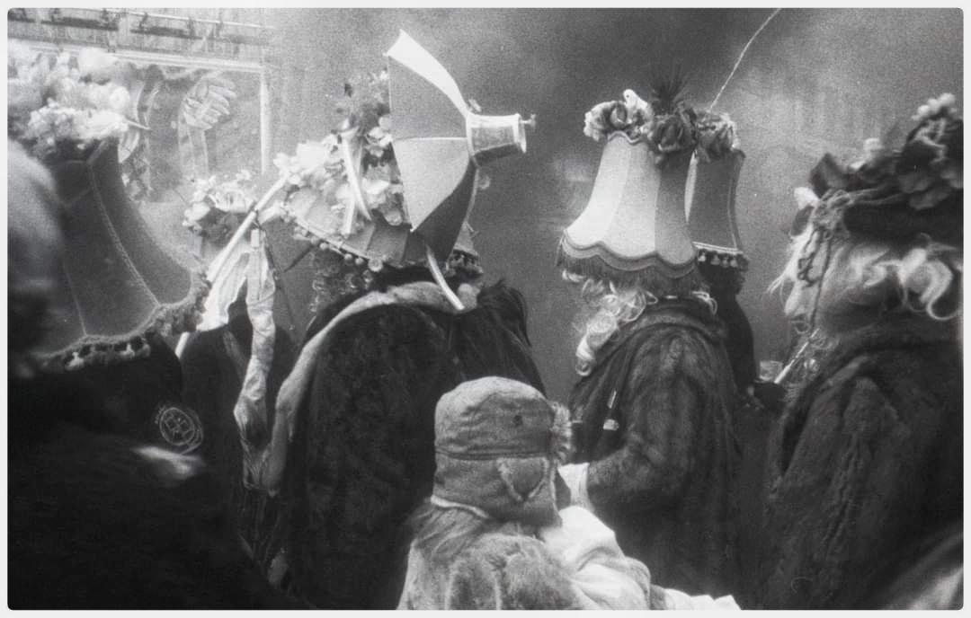 People in elaborate costumes with lampshades and umbrellas on their heads are gathered together.