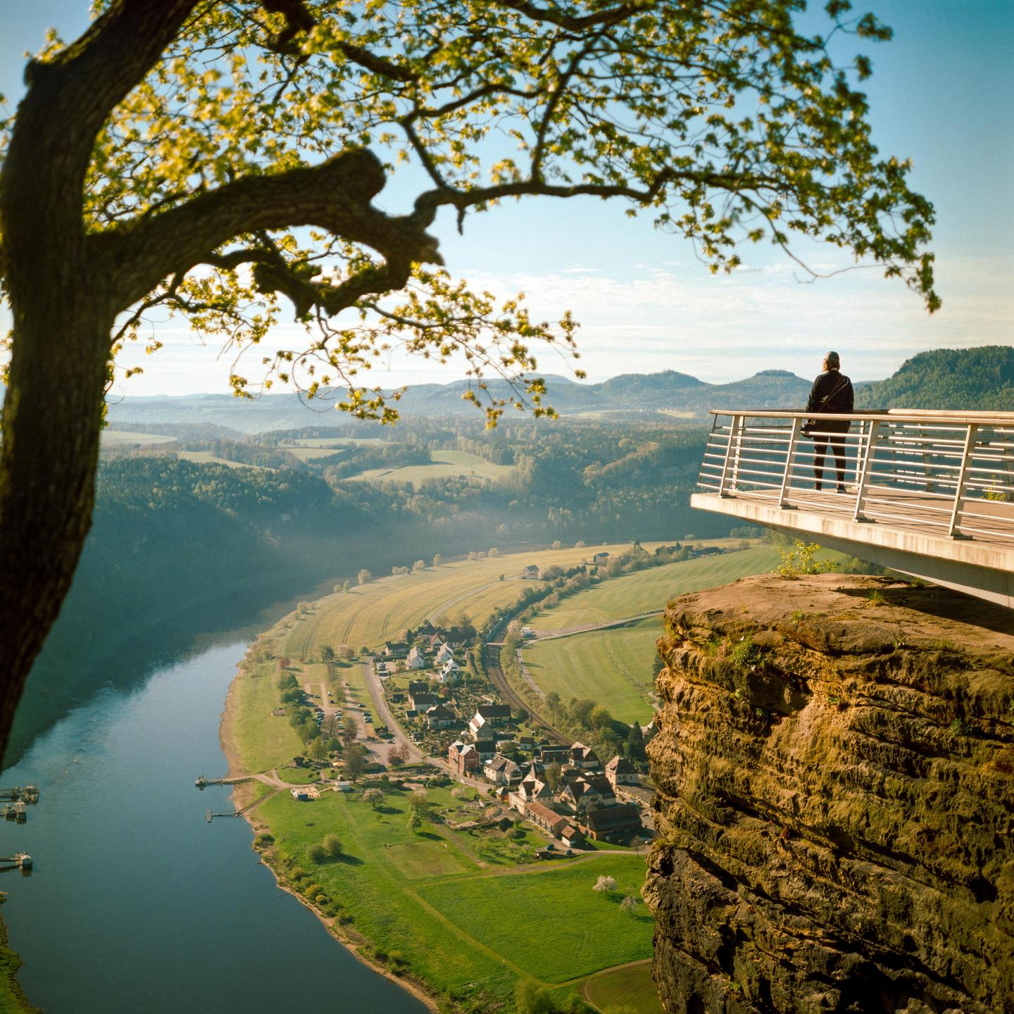 A person stands on a viewing platform overlooking a river, a village, and a valley with mountains in the distance, framed by a tree.