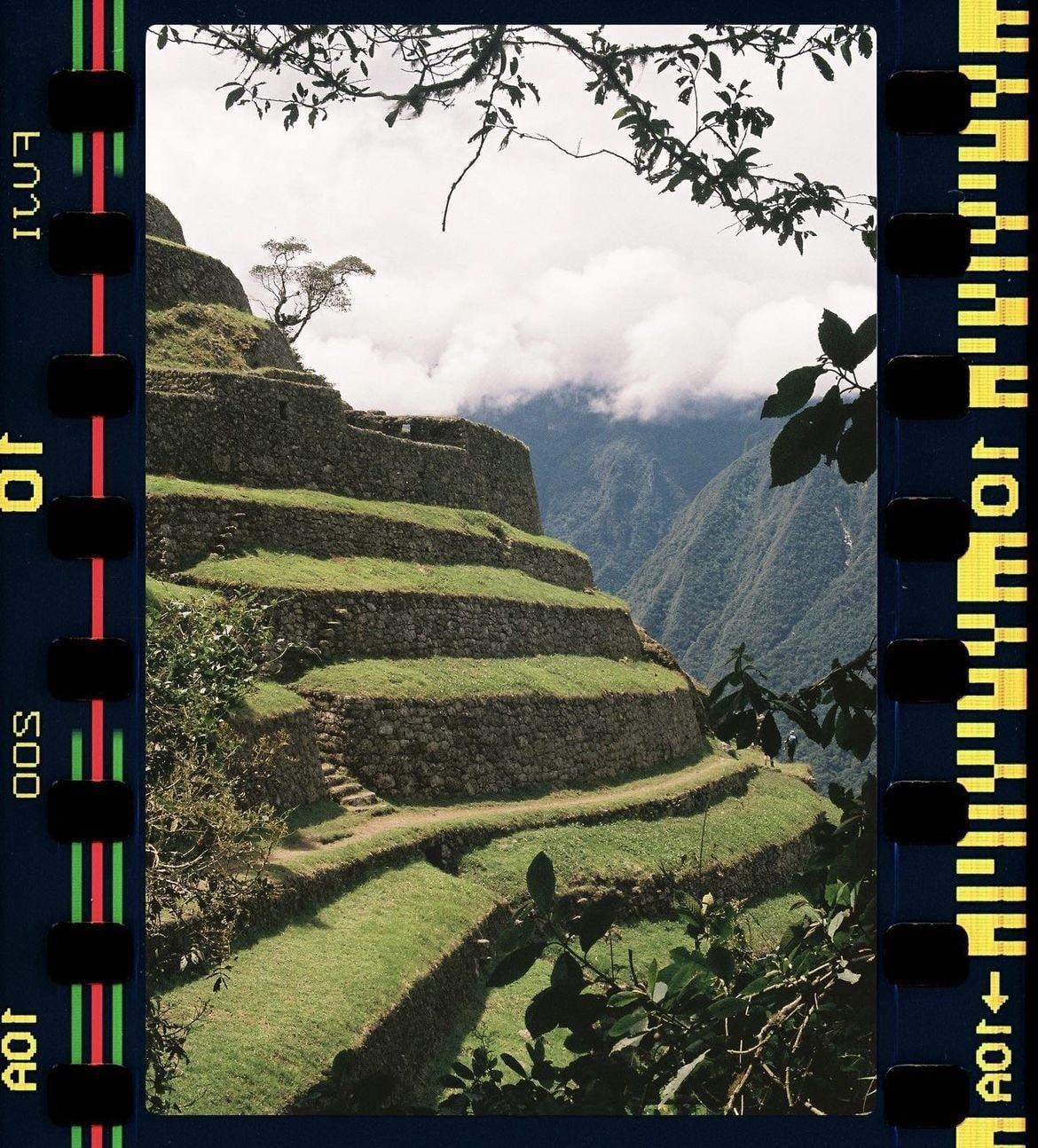 Ancient stone terraces covered in green grass ascend a hillside with mountains and clouds in the background.