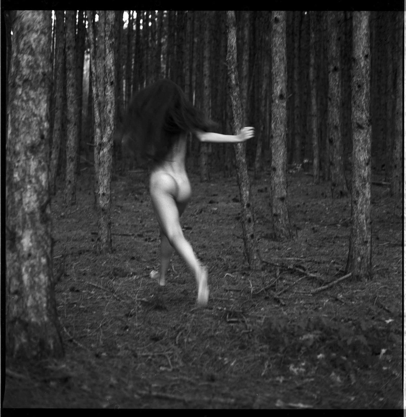 A nude woman with long hair runs through a forest of tall, slender trees.