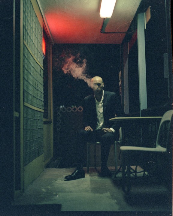 A bald man in a suit sits in a dimly lit hallway, exhaling a cloud of smoke.
