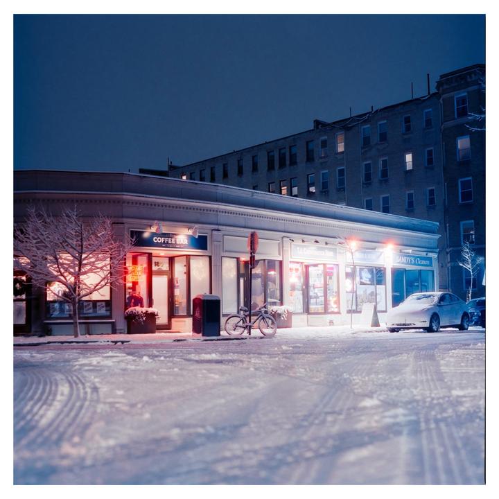 A snow-covered street with storefronts and a parked car is illuminated at night.