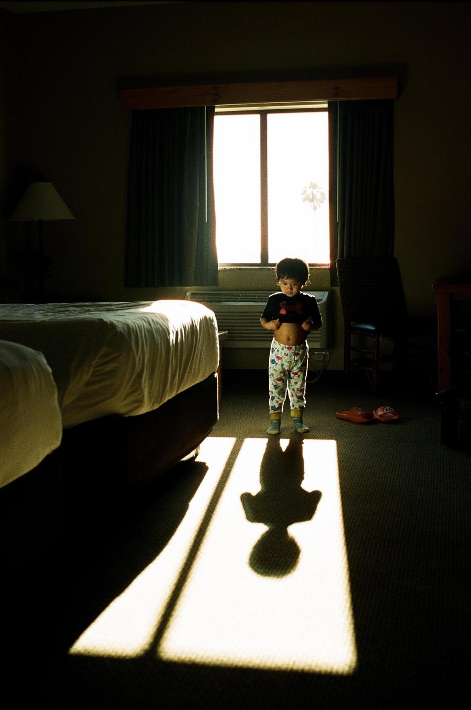 A child stands in a hotel room with a bright window behind them, casting a long shadow on the carpet.