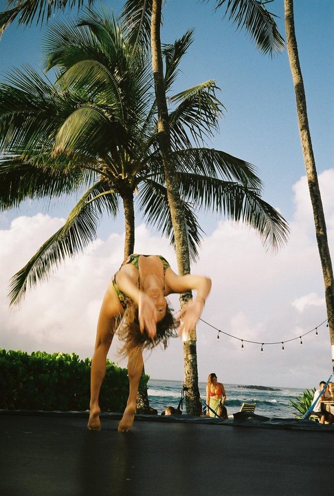A woman in a swimsuit is doing a backflip on a trampoline under palm trees with the ocean in the background.