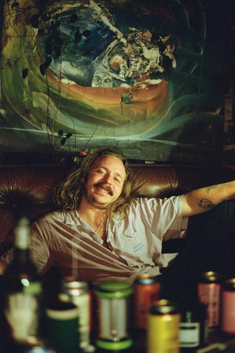 A man with long hair and a mustache smiles while sitting on a brown leather couch in front of a colorful abstract painting.