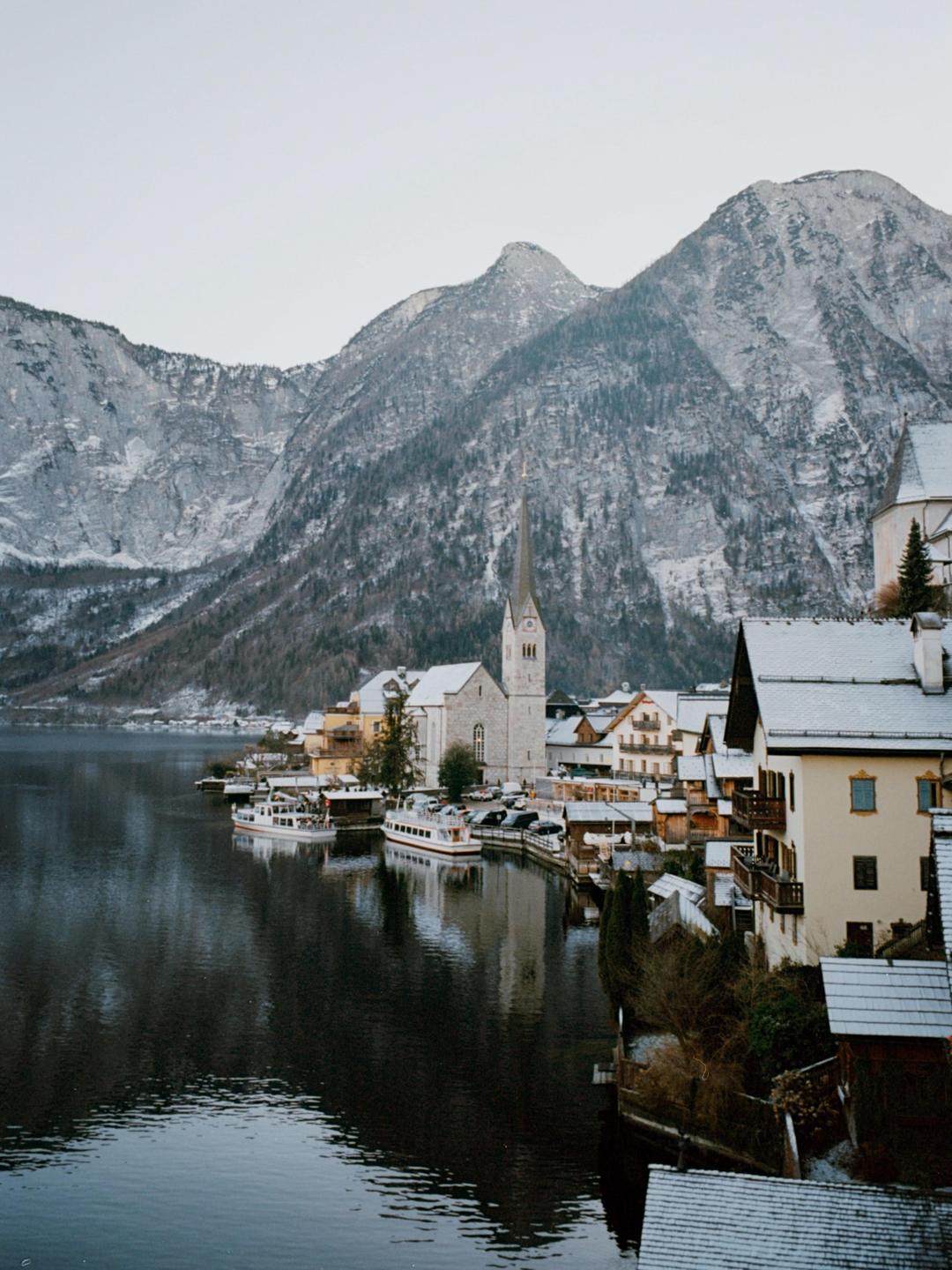 A town with a prominent church steeple is nestled beside a lake, surrounded by snow-dusted mountains.