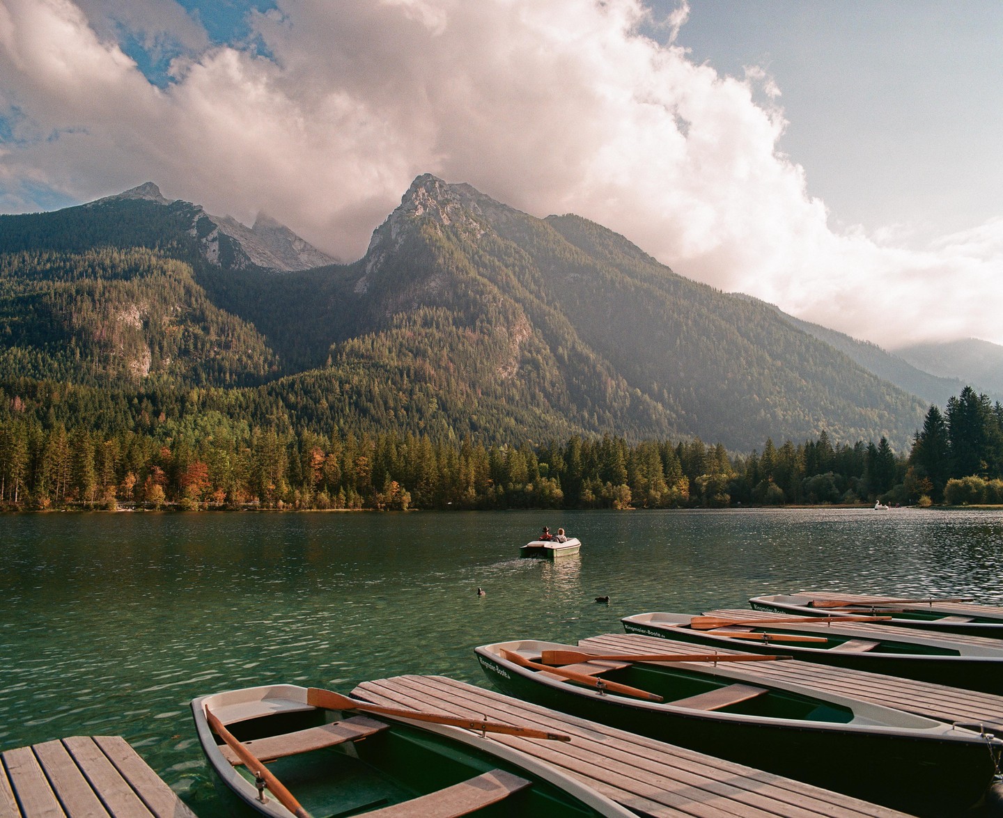 Several rowboats are docked on a lake with mountains and a forest in the background.
