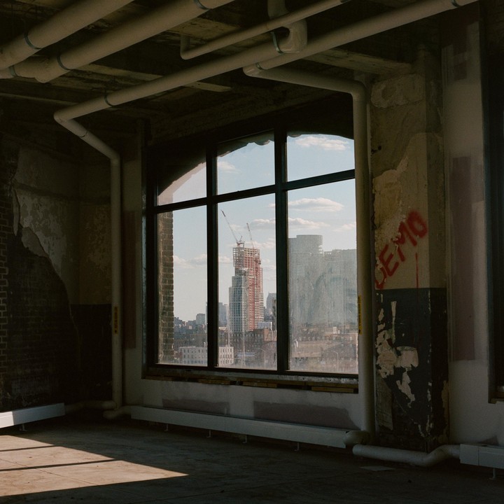 A city skyline with a building under construction is visible through a large window in a room with exposed pipes and peeling paint.