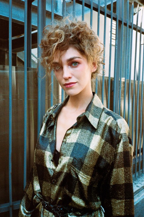 A woman with curly hair and a plaid shirt looks directly at the viewer.