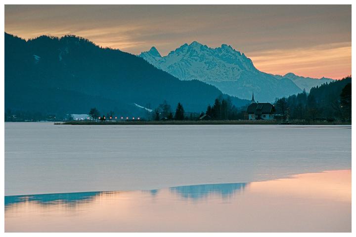 A frozen lake reflects the sunset sky, with mountains and a small village visible in the distance.