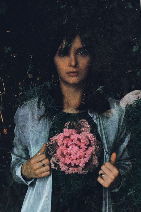 A woman with dark hair and a light shirt holds a large pink ornamental cabbage against a dark, leafy background.