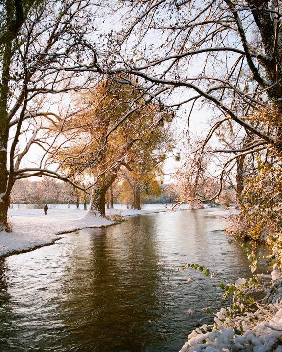A river flows through a snowy park with trees on both banks, and a person walks in the distance.