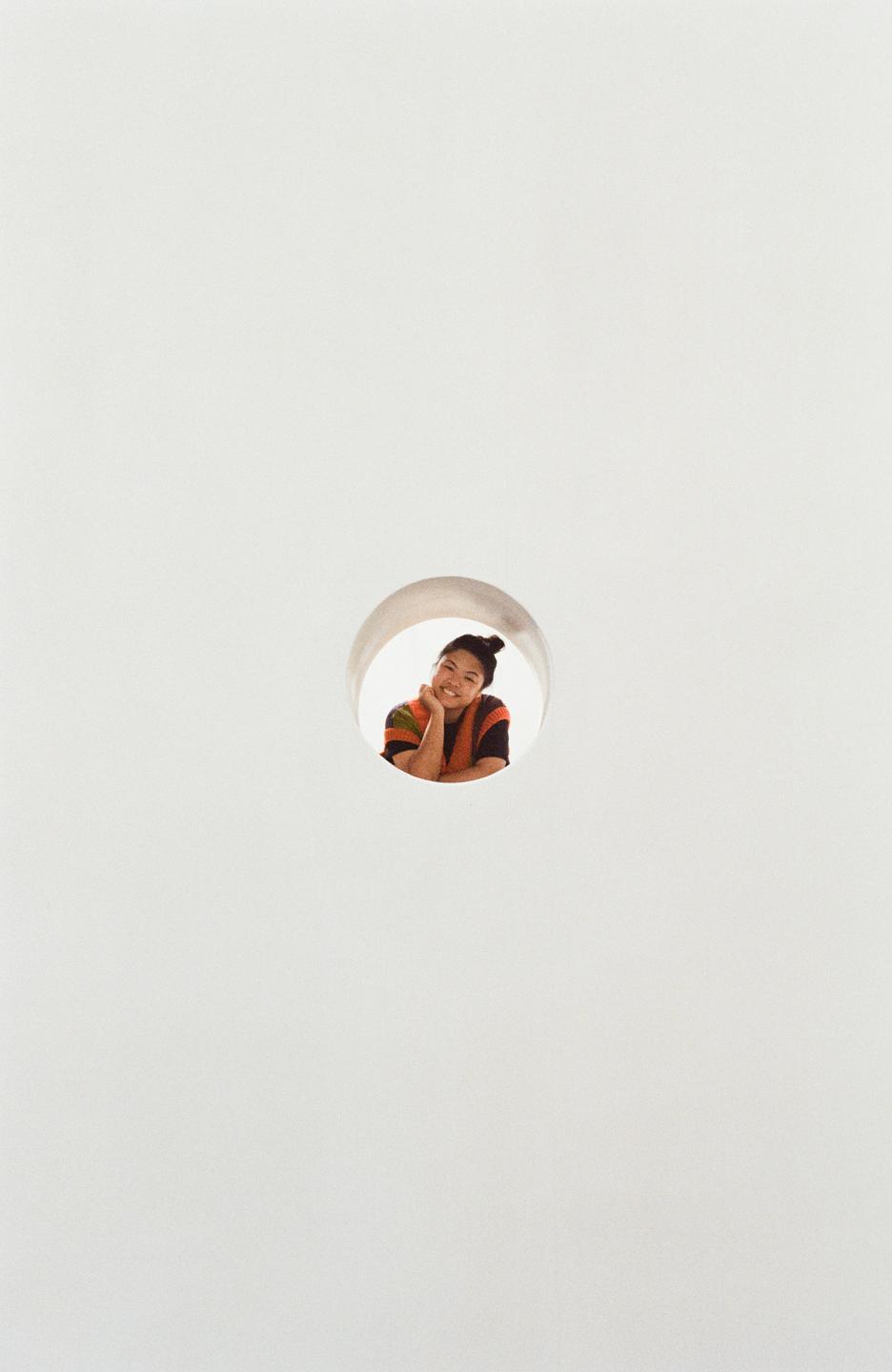 A woman with her hair in a bun smiles while looking through a circular opening in a white wall.