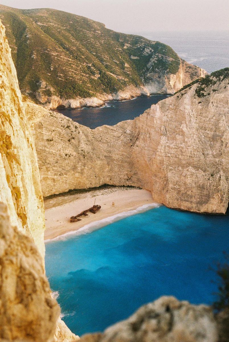 A shipwreck lies on a sandy beach surrounded by towering cliffs and turquoise water.