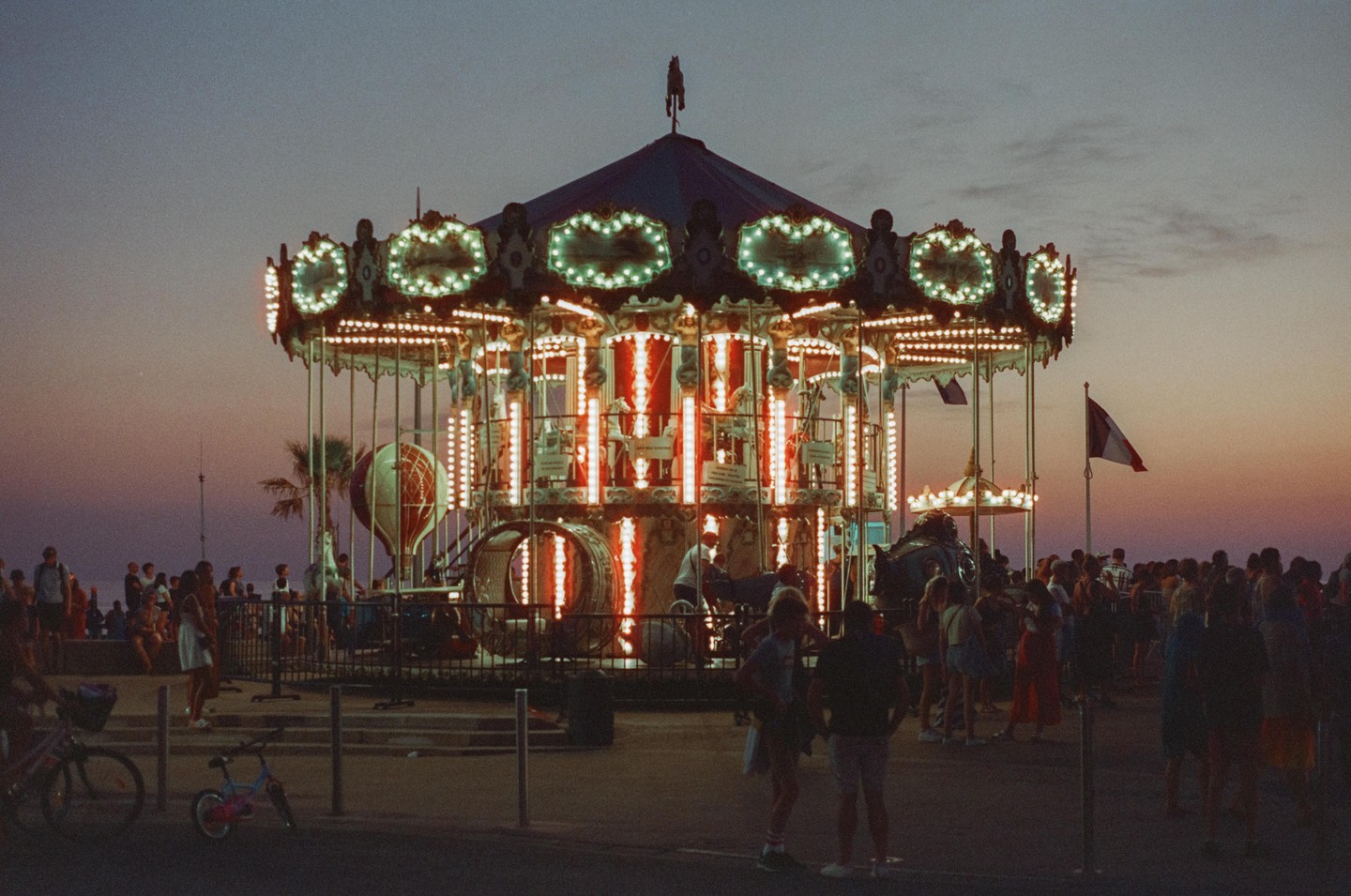 A brightly lit carousel stands at dusk, surrounded by people and a French flag waving in the background.