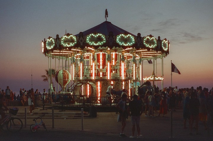 A brightly lit carousel stands at dusk, surrounded by people and a French flag waving in the background.