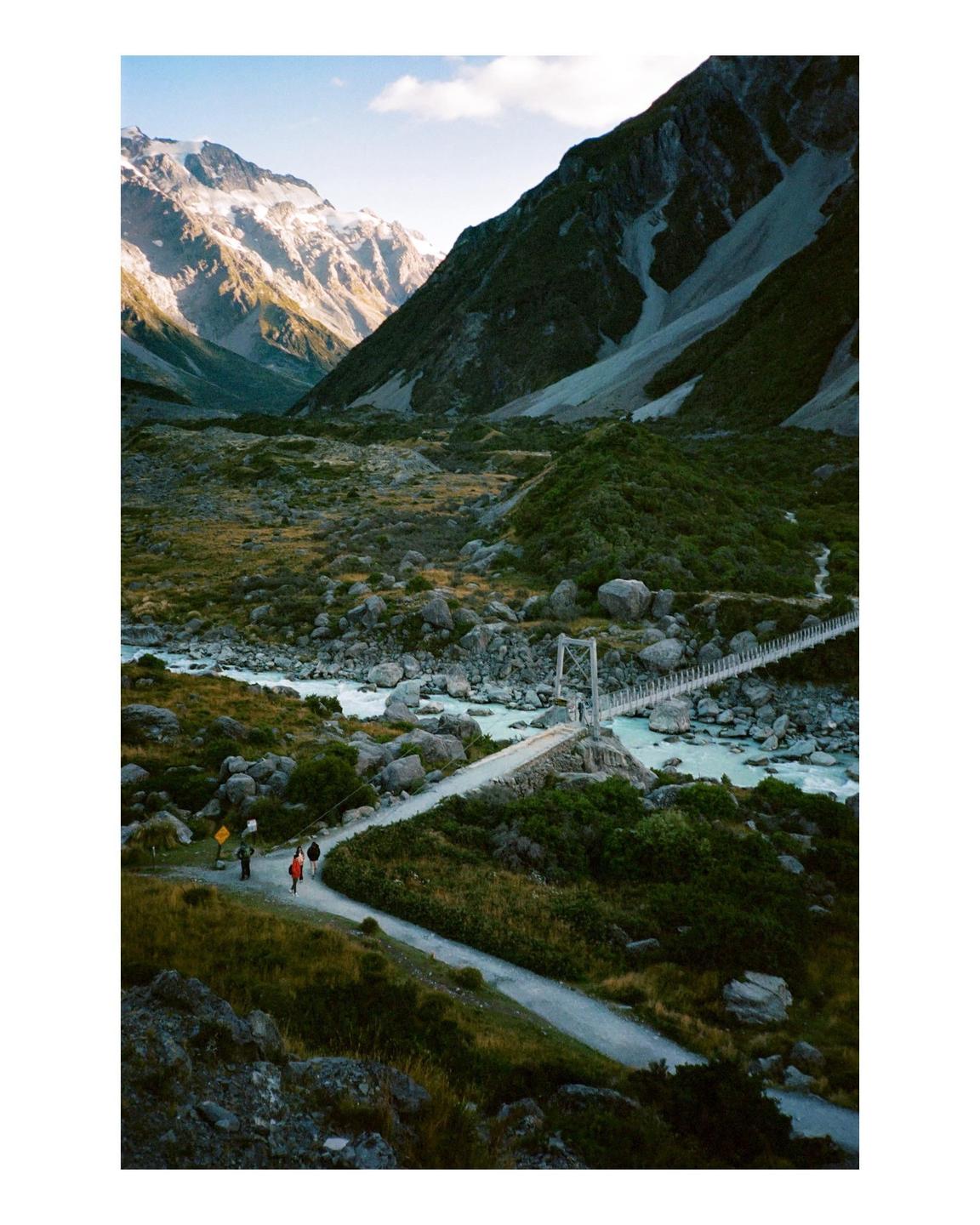 A group of people walk along a path towards a suspension bridge over a turquoise river, with snow-capped mountains in the background.