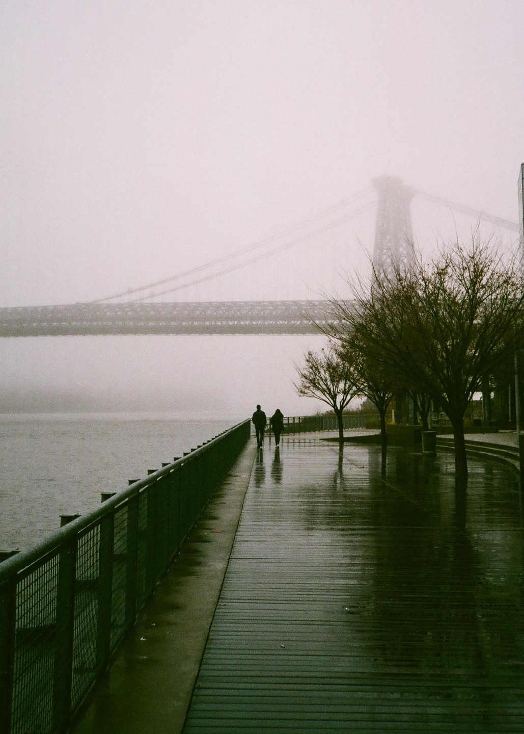 Two people walk along a wet boardwalk next to a body of water with a bridge in the background, partially obscured by fog.