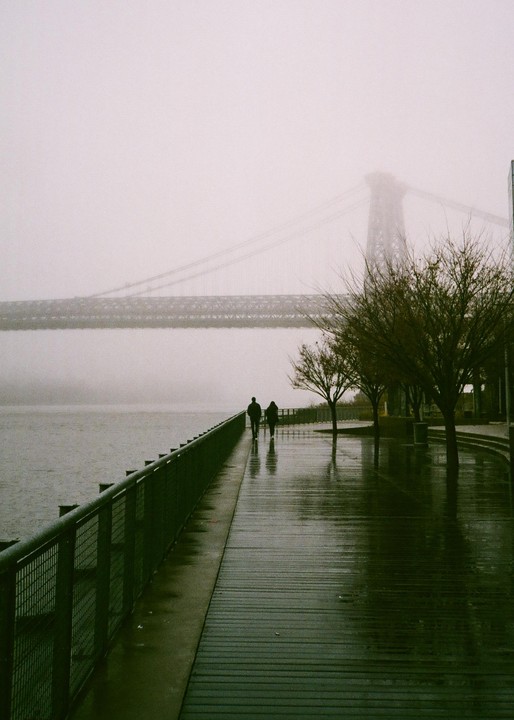 Two people walk along a wet boardwalk next to a body of water with a bridge in the background, partially obscured by fog.