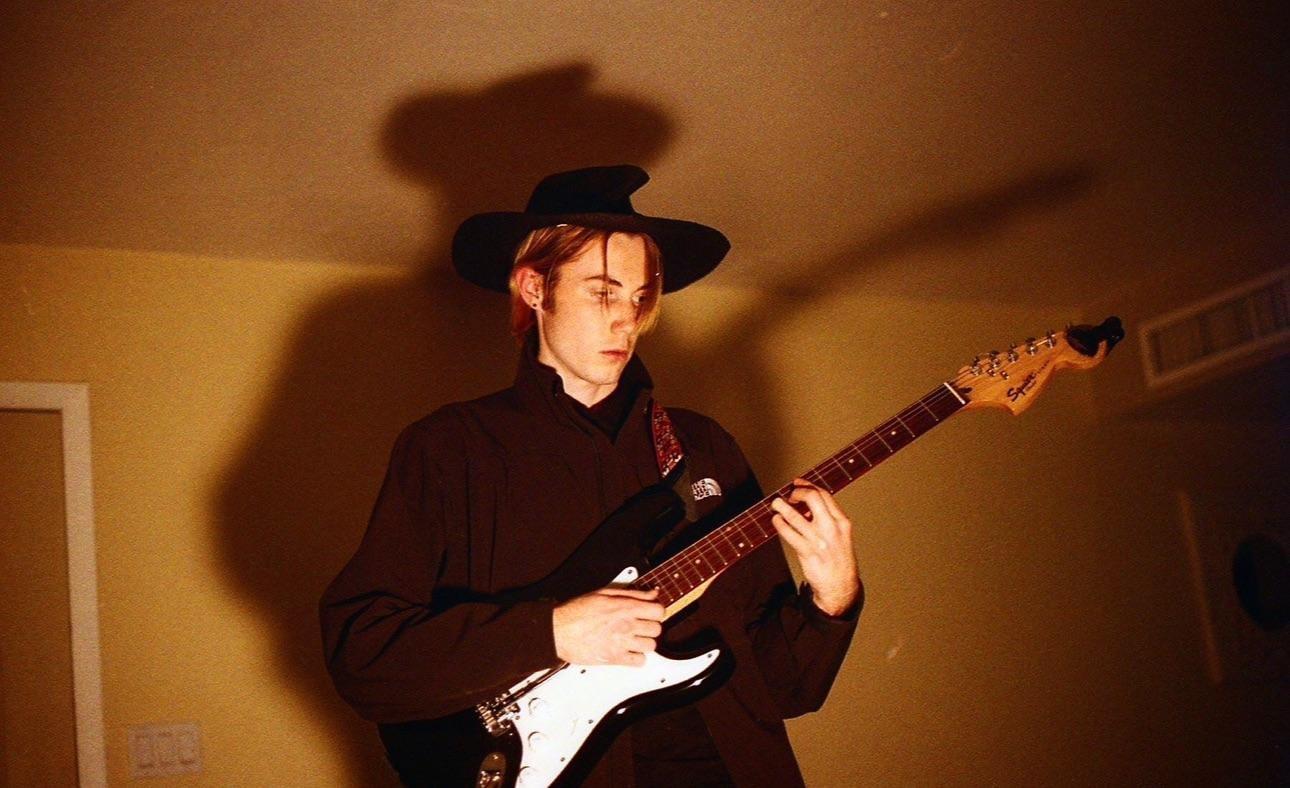 A young man wearing a hat plays an electric guitar indoors, casting a shadow on the wall behind him.