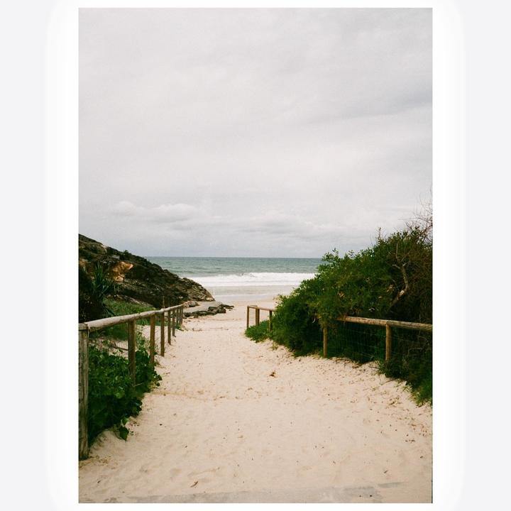 A sandy path leads to a beach with waves under a cloudy sky.