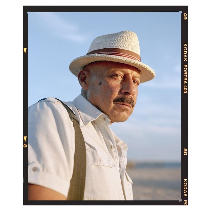A man wearing a straw hat and suspenders looks to the right with a serious expression.