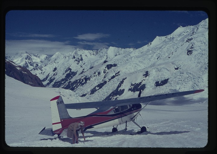 A man bends down next to a small plane on a snowy mountain with a clear blue sky.