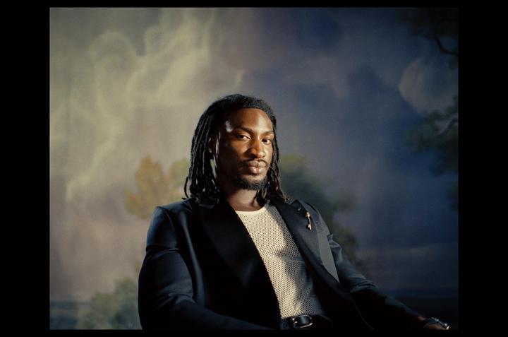 A man with dreadlocks and a beard is wearing a suit jacket and a mesh shirt against a painted backdrop.