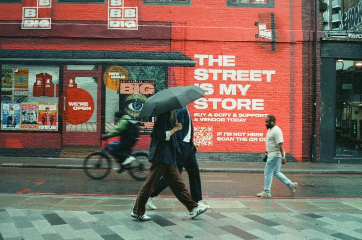 A person holding an umbrella walks past a red building with a mural that says "The Street is My Store" while a cyclist rides by.
