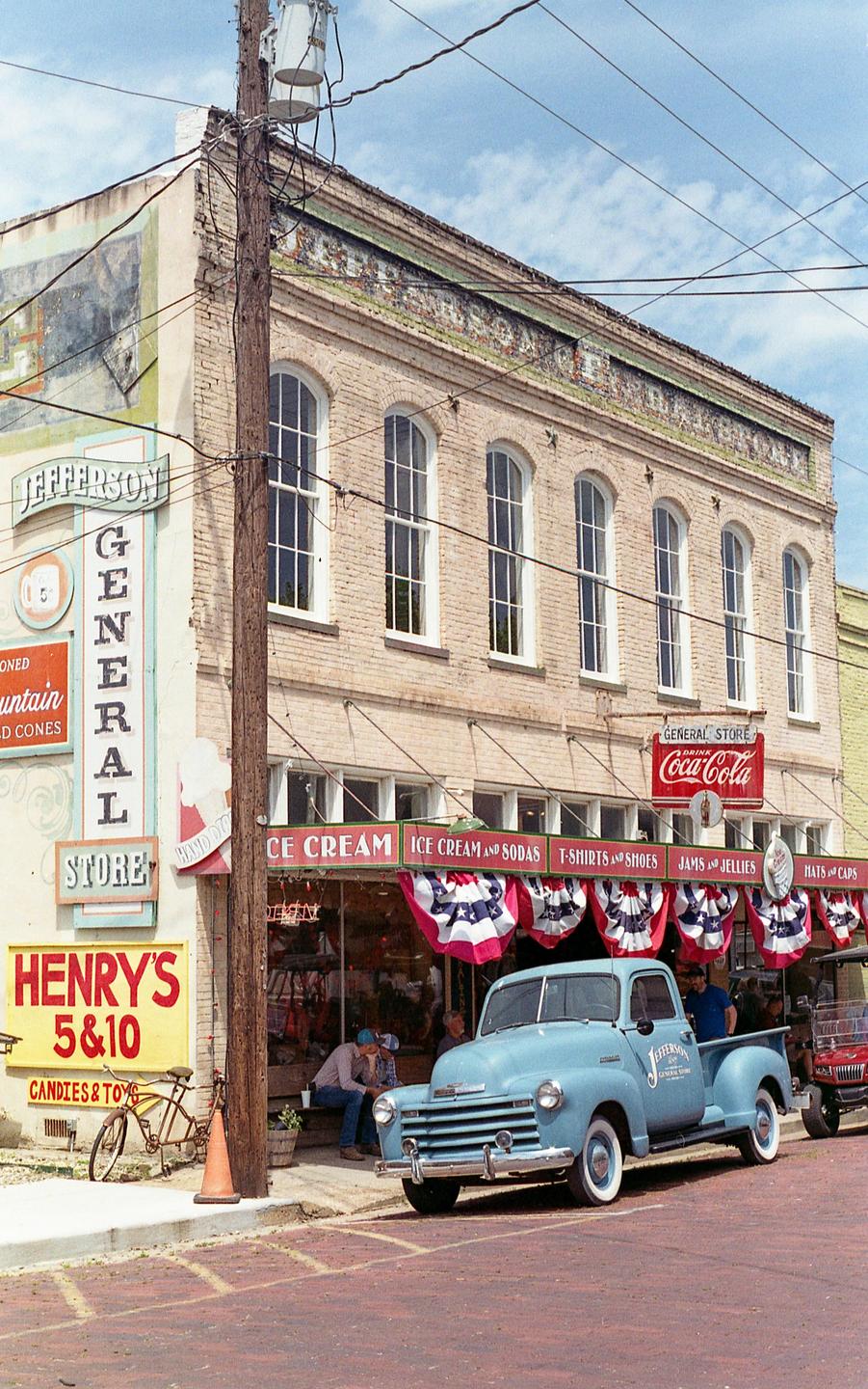 A light blue pickup truck is parked on a brick street in front of a general store with a red, white, and blue bunting.