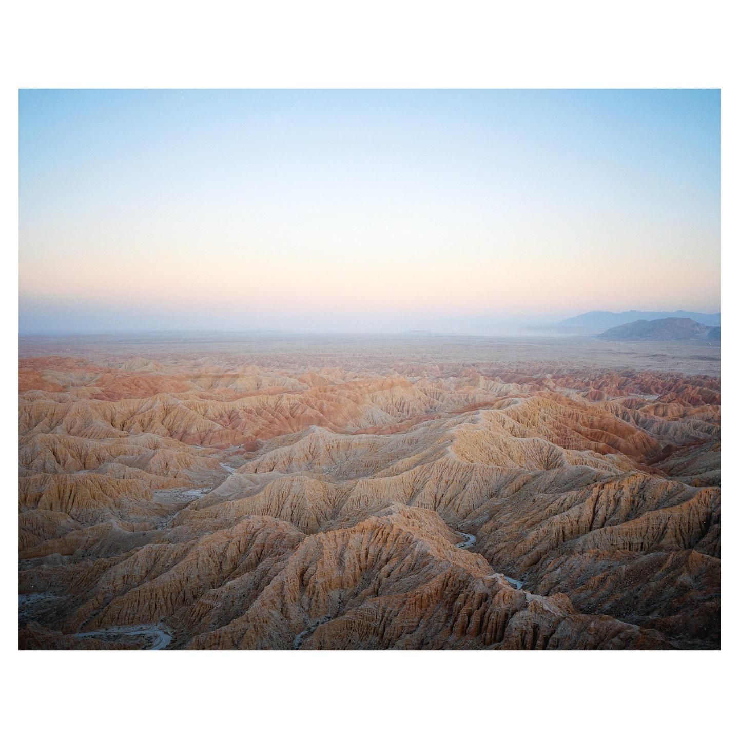 A vast desert landscape with intricate badlands stretches under a pastel sky at dusk.