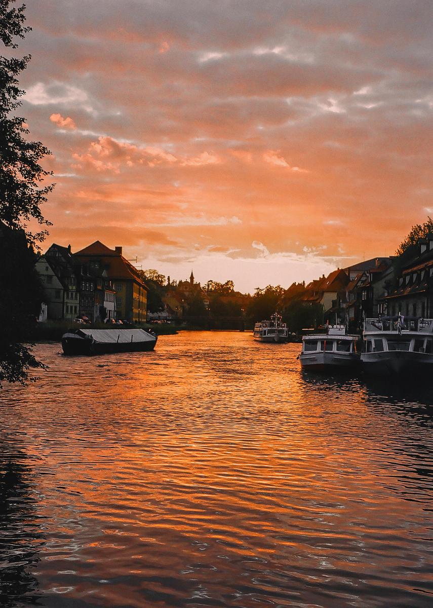 A river with boats and buildings on its banks is illuminated by the orange light of sunset.