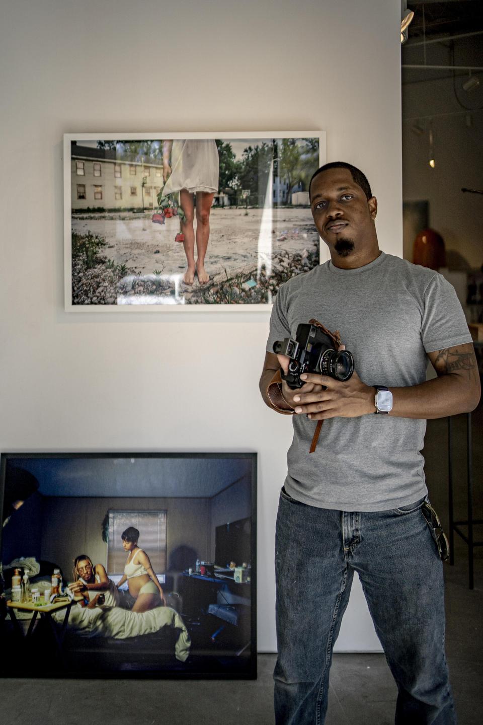 A man stands in a gallery holding a camera, with two framed prints on the wall behind him.