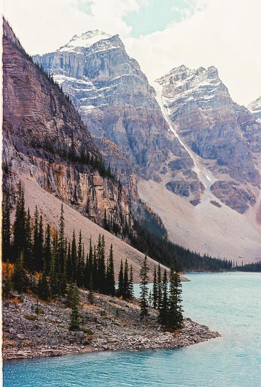 A turquoise lake is surrounded by a rocky shore with pine trees and large mountains with snow-capped peaks.