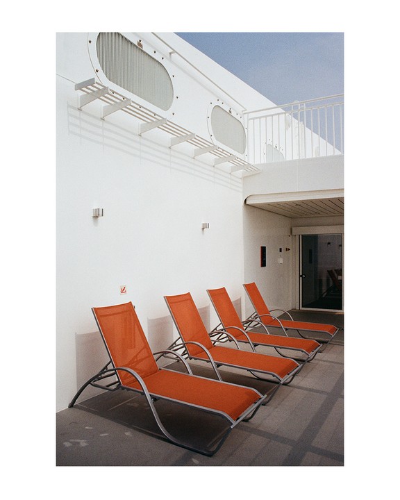 Five orange lounge chairs are lined up against a white building with oval windows.