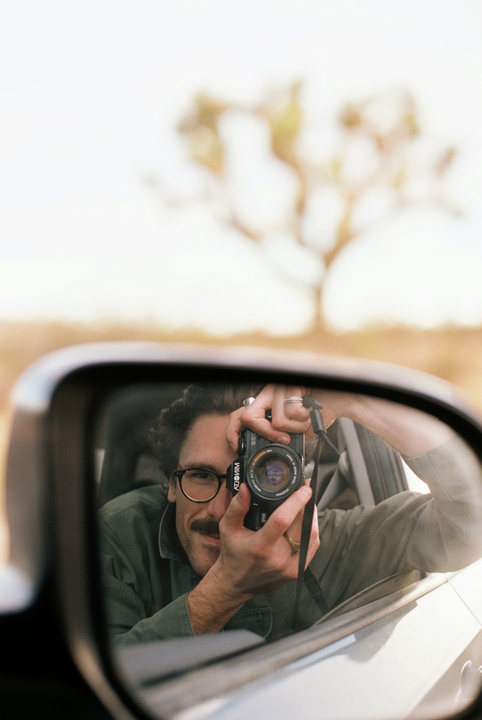A man with a mustache and glasses takes a self-portrait in a car's side mirror, holding a camera to his face.