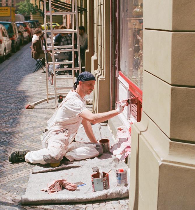 A man wearing a bandana and paint-splattered clothes paints a window frame red on a cobblestone street.