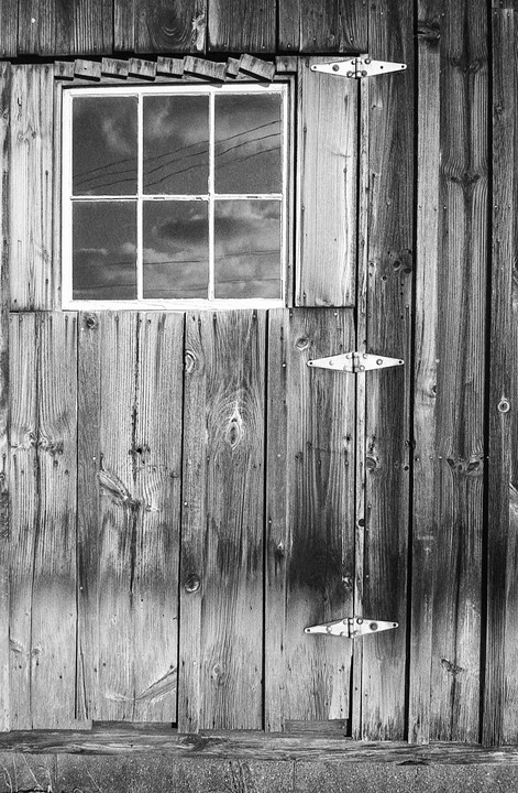 A window with a reflection of clouds and power lines is set into a weathered wooden door with three hinges.